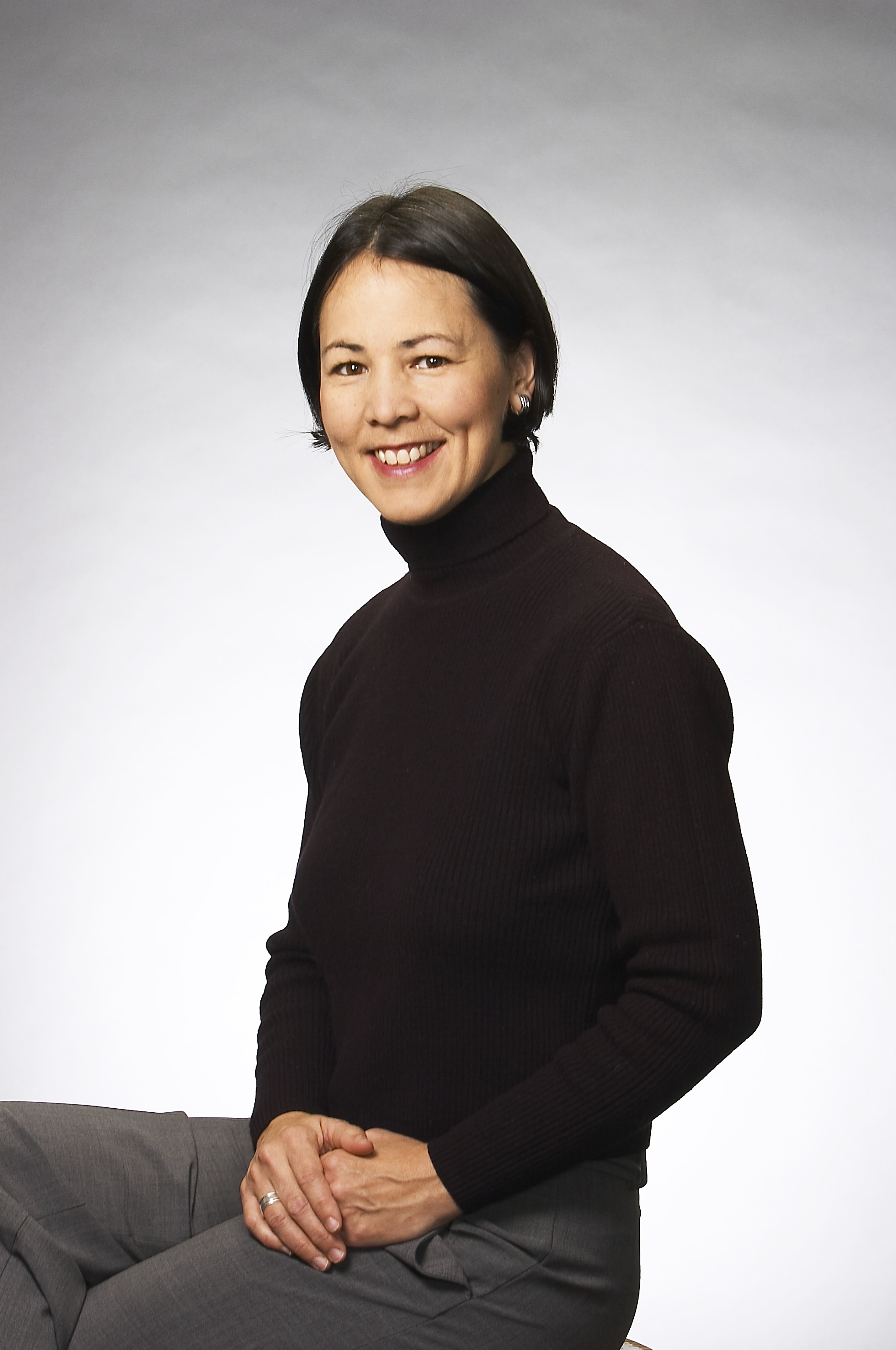 Radio announcer May-Lily Lee sitting on a stool in a pose. She is wearing a long-sleeved black turtle neck and gray pants. She is smiling and her hands are resting one atop the other on her leg.