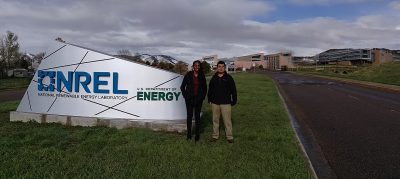 Two students stand in front of a sign with a large laboratory complex in the background. 