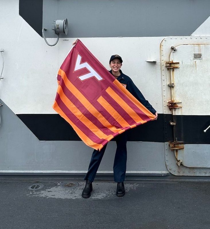 Alexandra Scarfe smiles while holding a striped Virginia Tech flag on a U.S. Navy ship. She is in her Navy uniform smiling for the camera.