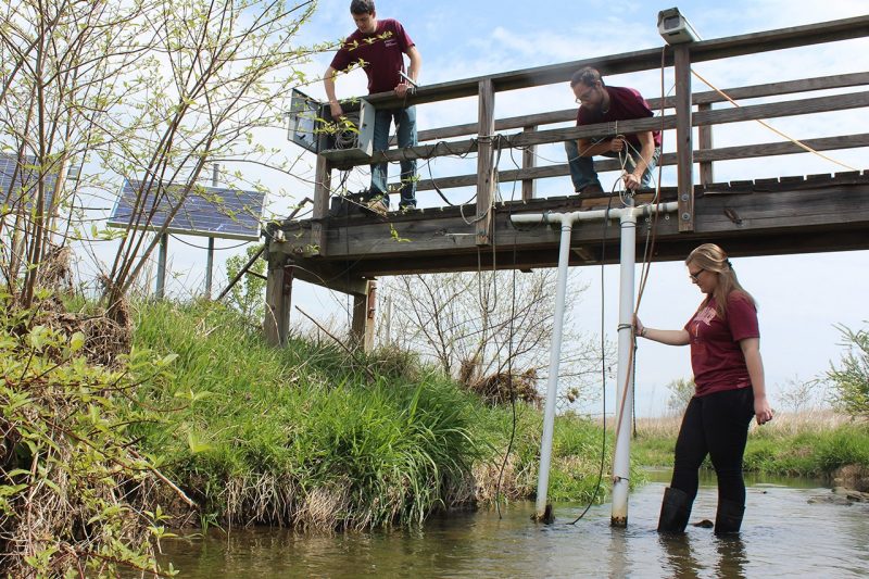 Professor and students stand on a bridge and in the water at Stroubles Creek