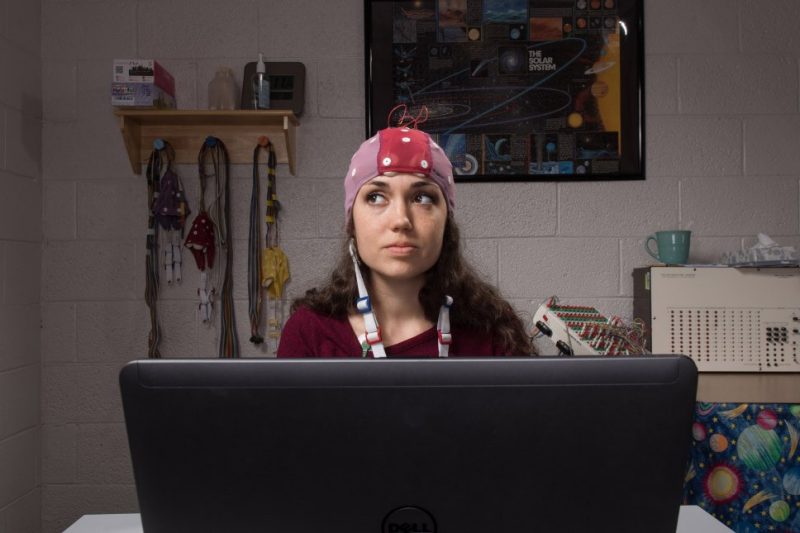 In the photo, Sarah Hanson wears a cap covered with electrodes while sitting in front of a laptop.