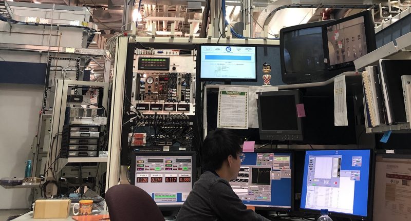 Chemistry graduate student Zhijie Yang is operating synchrotron measurement computer at Advanced Photon Source of the Argonne National Lab in a photo taken before the COVID-19 pandemic. A bay of computer monitors surround him. 