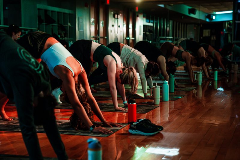 The photo takes place in a dimly lit gym with a shiny, reflective wooden floor and a wall of windows in the back. Several students in athletic wear are in a line, all doing a downward dog yoga position. Each has a mat under them and water bottles in front of them as well.