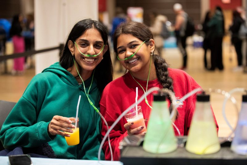 Two female students in hoodies wear small colored breathing tubes, hooked up to a machine with different types of scented air. They both hold clear plastic cups with smoothies. Several out of focus students in the background are standing in a line.