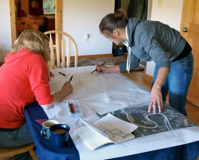 Two female college students work at a table with renderings and maps. 