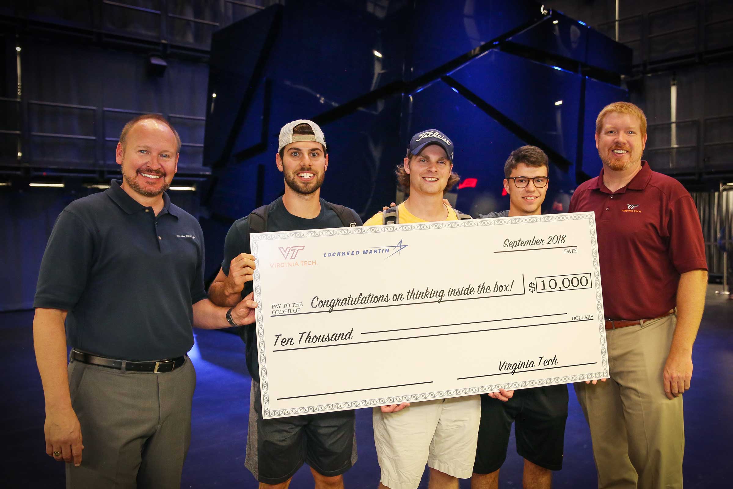 Three male, senior engineering students stand smiling holding a large check that says congratulations on thinking inside the box.