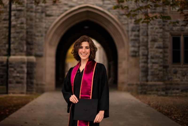 Abbey Stone stands in front of a building on the Virginia Tech campus wearing her graduation gown.