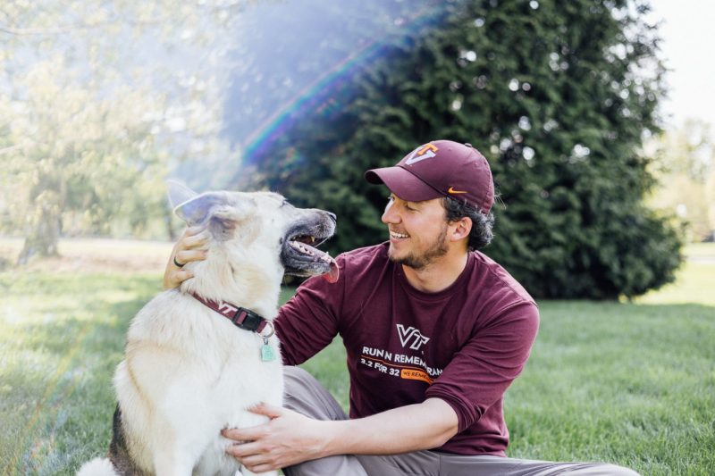 A German shepherd looks at her owner, Seth Bagbey, a young man wearing a maroon Virginia Tech t-shirt and maroon VT hat.