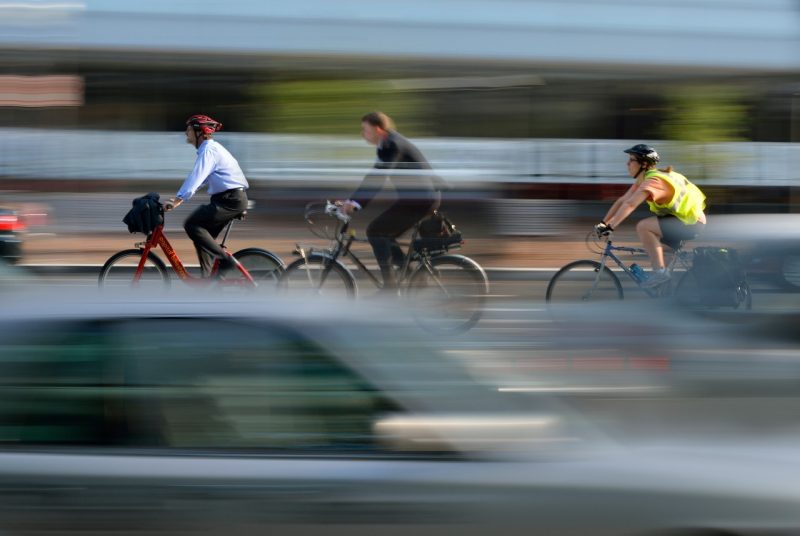 Three people on bicycles travel through Washington, D.C. as traffic rushes by. 