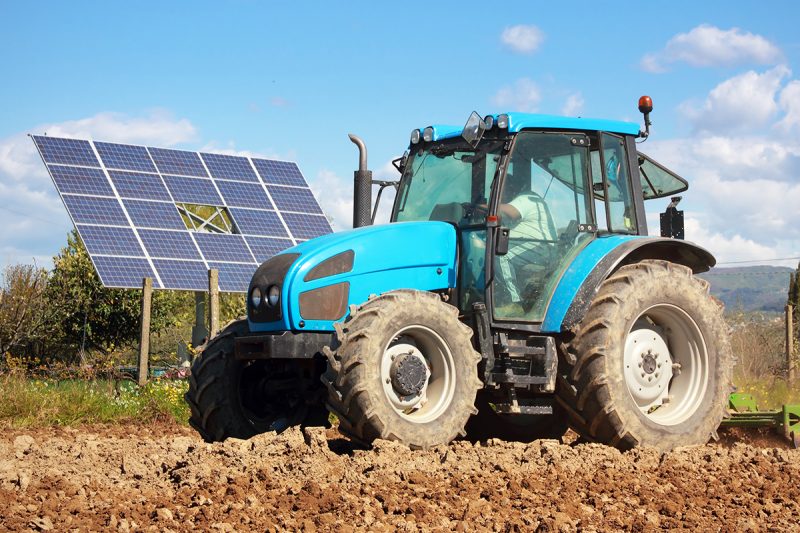 A tractor sits in a plowed field next to a solar array. 