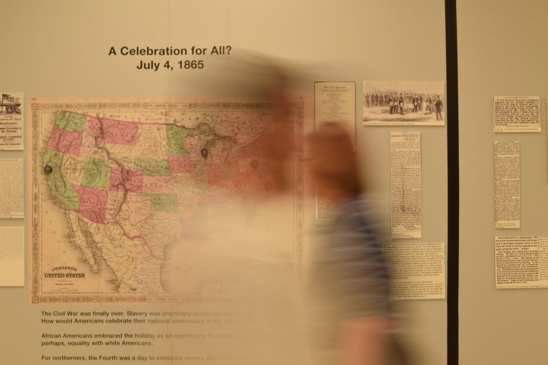 Two blurred images of people in front of a poster at the Mapping the Fourth of July exhibit.