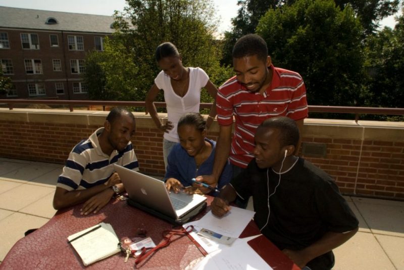 A group of Haitian students are gathered around a computer on an outdoor patio.