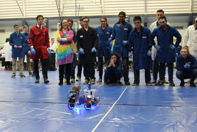 The Chem-E Car team is standing with blue lab coats on and watching their car roll in front of them.