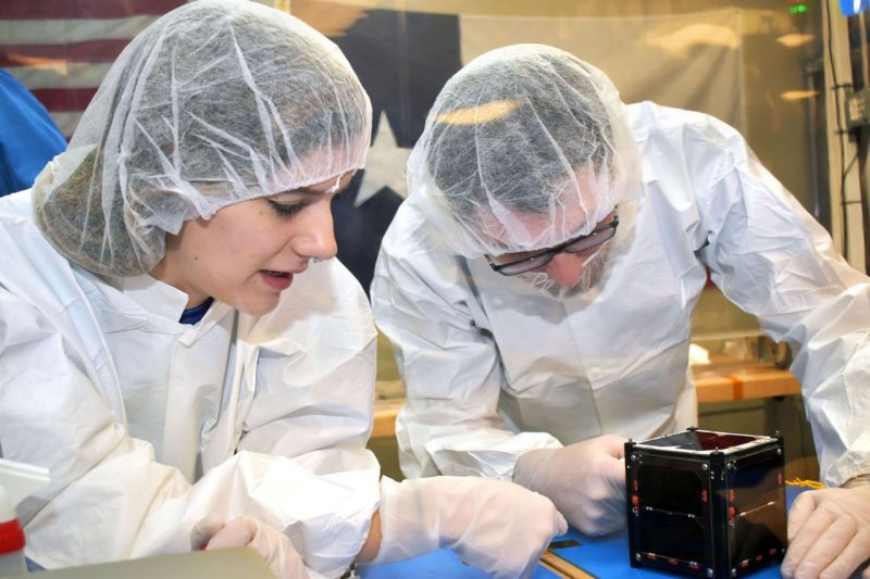 Two students wearing protective clothes and hair nets work on a satellite.