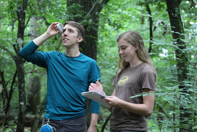 A young man and young woman stand in a forest. The man looks up while holding a clinometer to one eye. The woman looks down at a clipboard she is holding.