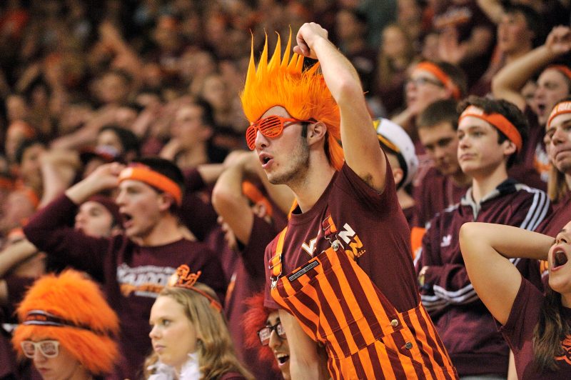 A fan wearing orange and maroon striped bibs and an orange spikey-haired wig cheers in the student section at Cassell Coliseum