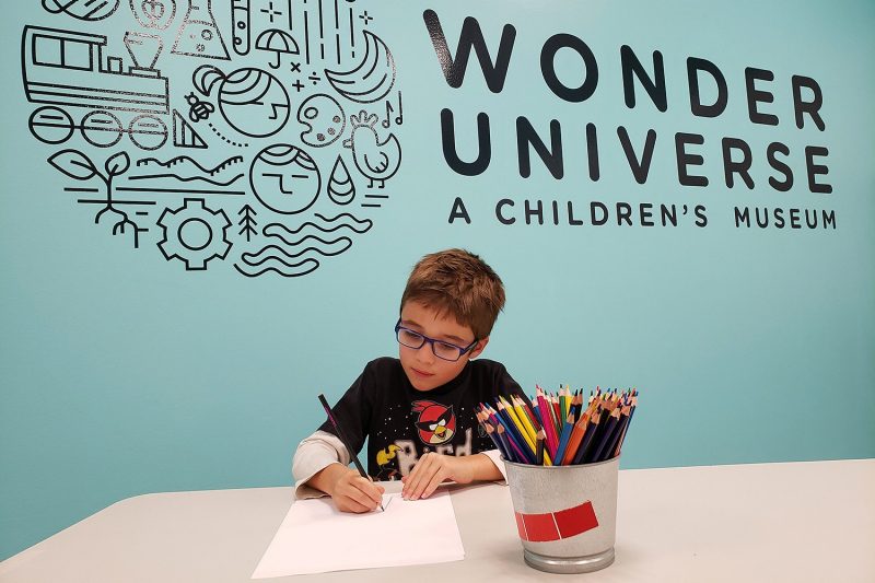 A young boy draws with colored pencils in front of a blue wall with the logo for Wonder Universe.