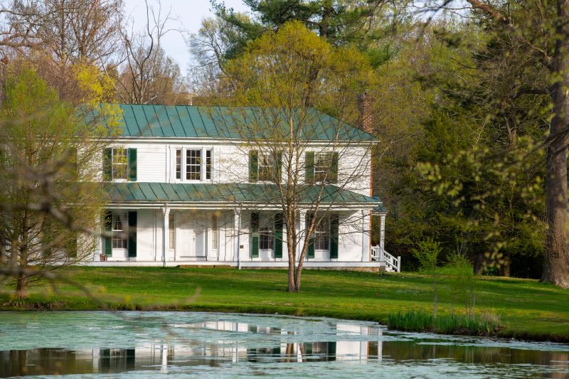 The White Solitude House with green shutters sits behind the glassy Duck Pond on a clear, spring day.