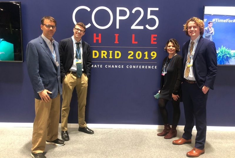 Three men and one woman standing in front of a large wall hanging for COP25.