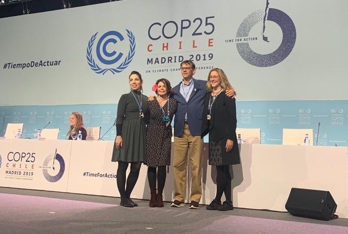Three women and one man standing together in front of a long table with a large COP25 banner hung behind it.