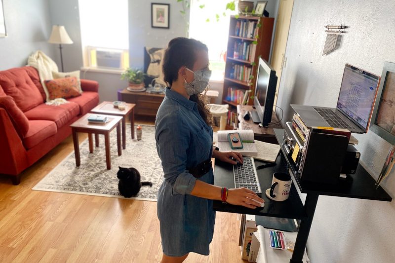 Whitney Hayes stands at her home office desk, while her black cat joins her.