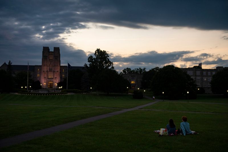 Two people sit with their back to the camera having a picnic, facing Burruss Hall, on the Drillfield at dusk.