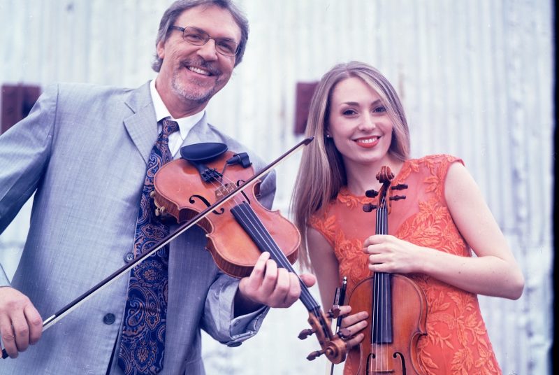 Mark and Maggie O'Connor pose outside in front of a barn holding their fiddles.