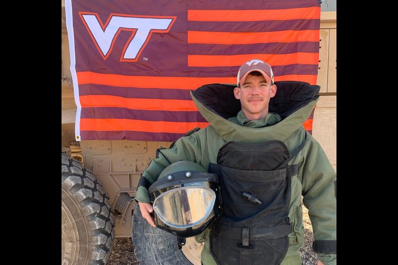 U.S. Army Sgt. Alexander Unkle, explosive ordnance disposal technician, stands in protective gear in front of a Virginia Tech flag.