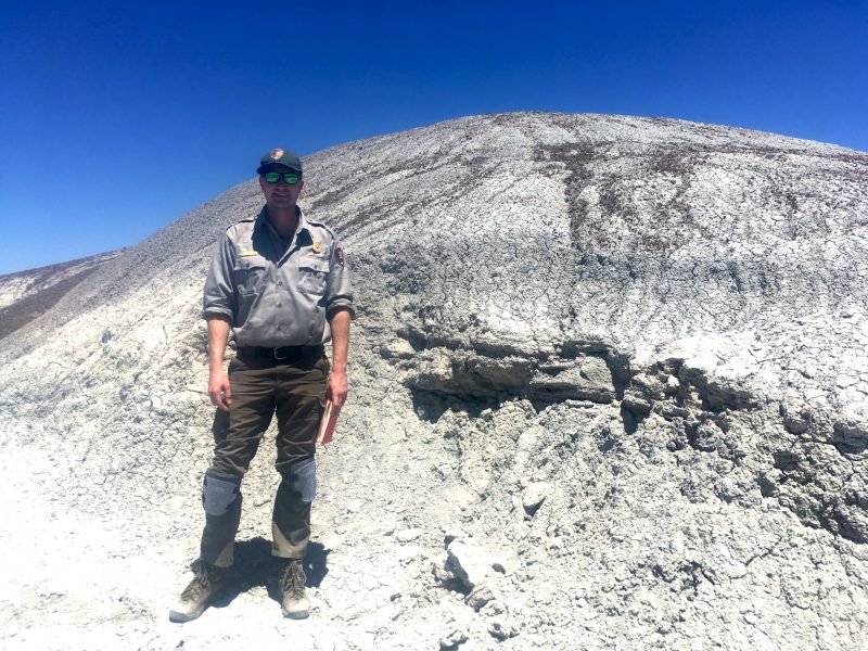 Ben Kligman poses at Arizona's Petrified National Forrest Park in 2019, where he worked as a seasonal paleontologist. Ben is wearing a park ranger's uniform, while standing in front of massive ancient rocks. Photo by Adam Marsh.