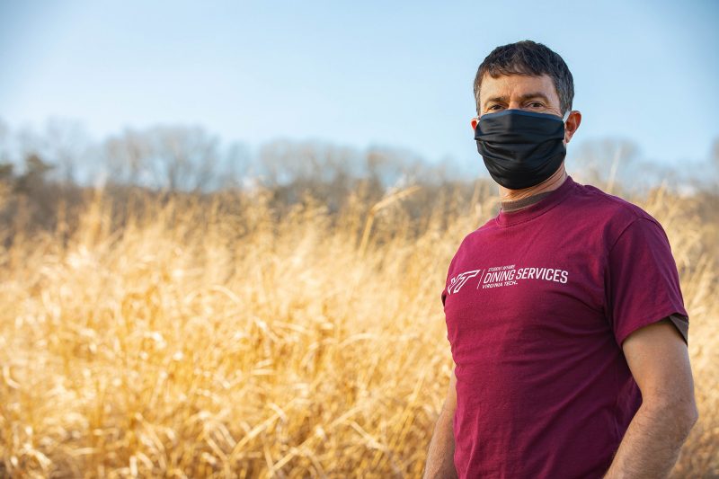 Jadrnicek standing in Virginia Tech shirt, wearing a face covering in front of a field of wheat