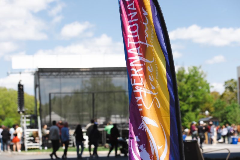 A rainbow-colored blade-shaped flag ripples in the breeze. It has "International Street Fair" and "Sponsored by the Cranwell Family" written in white with a globe icon. Several people and a large stage are out of focus in the background.